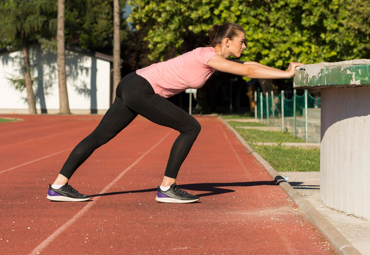 на біговому треці, розташованому у спортивному комплексі стоїть жінка, та вионує розтягнення м'язів