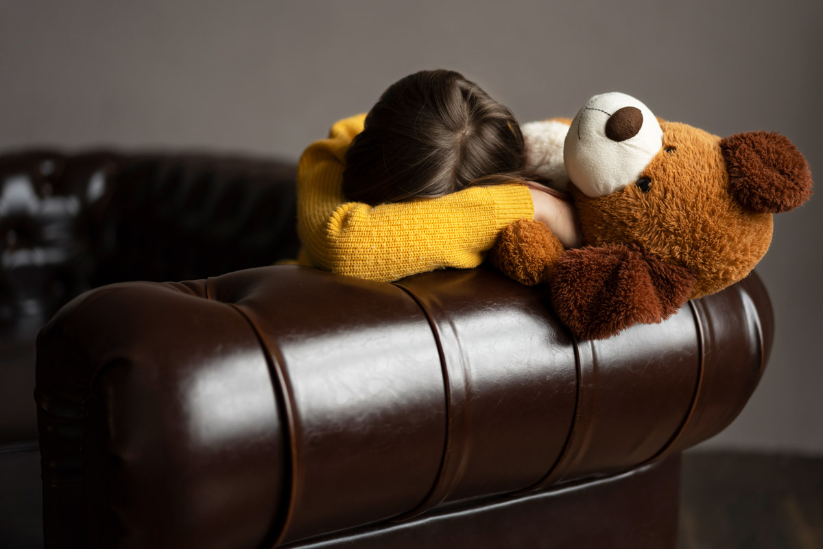 замсучена дитина сидить з іграшковим ведмедиком та ховається на дивані за своєю рукою