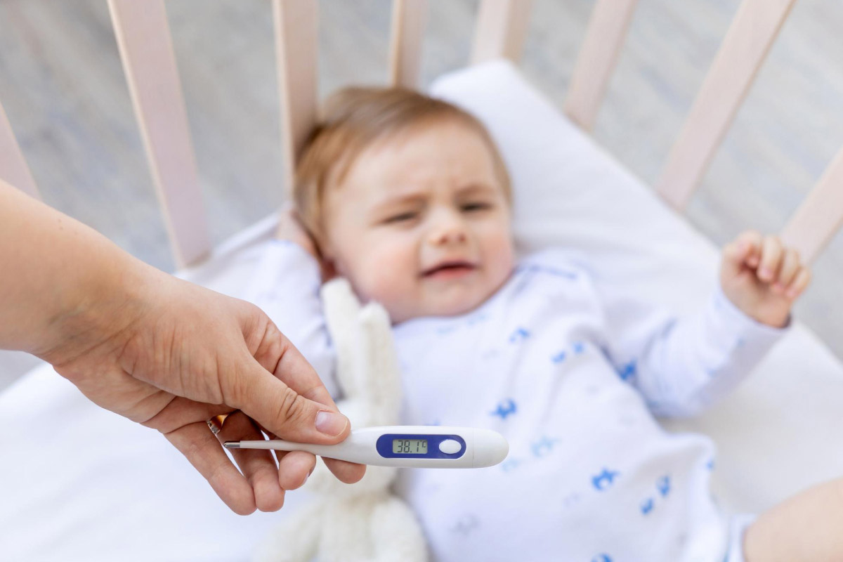 дитина лежить в колисці та плаче, а в руці мами знаходиться термометр