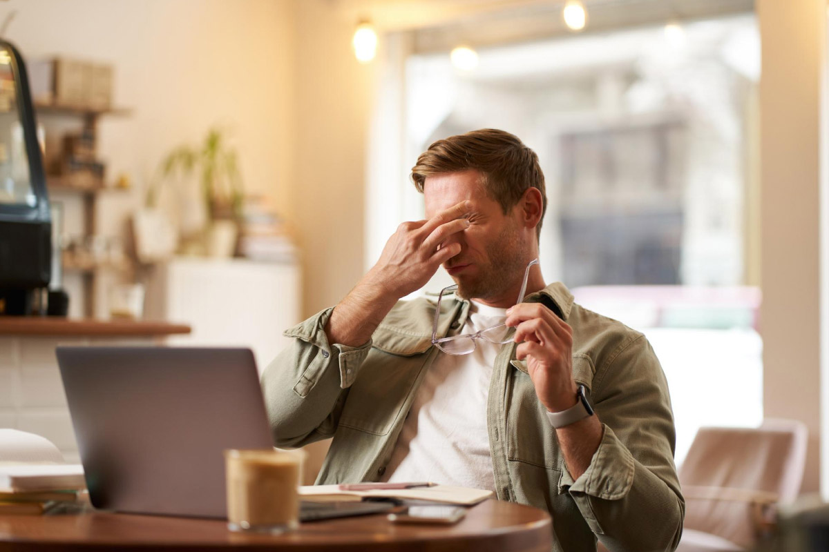 втомлений чоловік сидить за робочим столом, працює за ноутбуком та потирає очі через втому