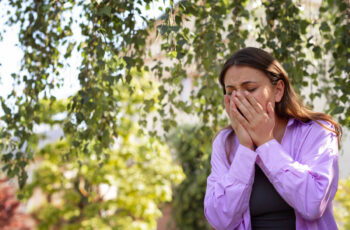 жінка на вулиці, теплим літнім днем, стоїть під березою, та прикриває обличчя руками, вочевидь, її турбує кашель та чхання, які викликані алергією