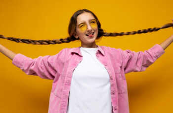 дівчинка на жовтому фоні в рожевій сорочці з довгими косами, які вона тримає в руках, посміхається та виглядає щасливою