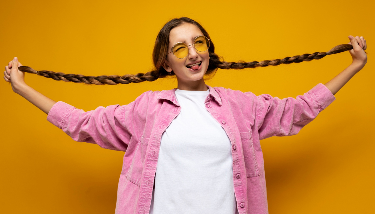 дівчинка на жовтому фоні в рожевій сорочці з довгими косами, які вона тримає в руках, посміхається та виглядає щасливою