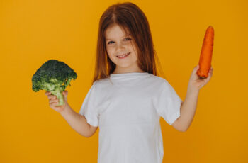 зображення дівчинки на теплому помаранчевому фоні, вона тримає в ркках овочі та посміхається