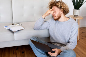 втомлений чоловік тримає ноутбук та сидить на підлозі в апатії