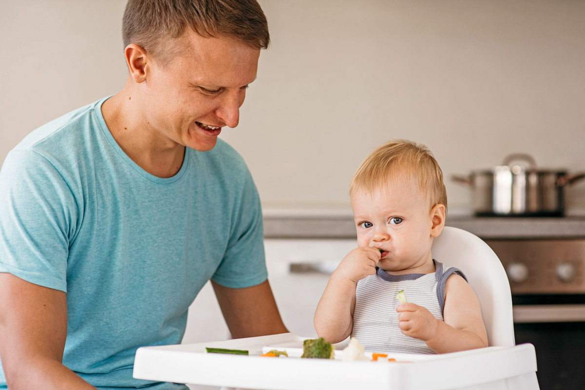 дитина сидить в дитячому кріслі та їсть, за нею дивиться батько